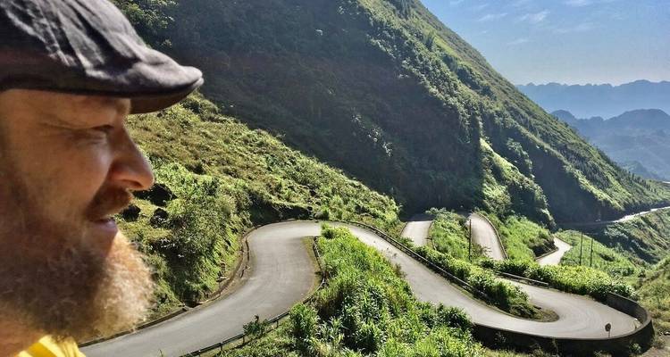 Nahaufnahme-Profil eines Reisenden, der auf einen gewundenen Bergpass blickt, der sich durch steile grüne Hänge schlängelt.
