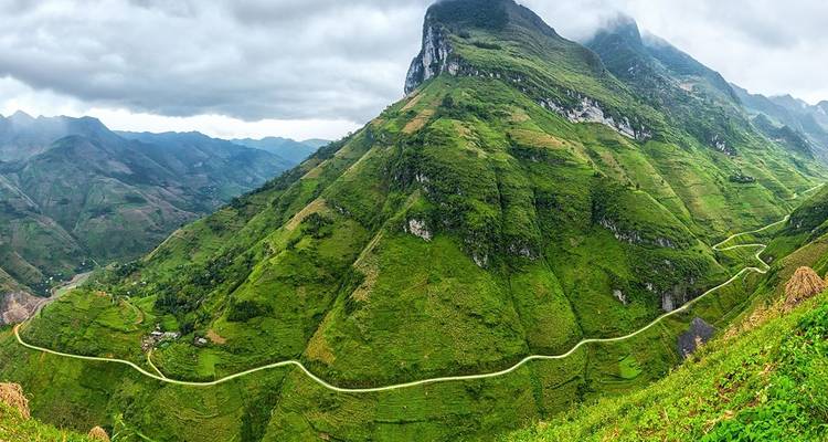 Panoramablick auf einen hoch aufragenden grünen Berg mit einer sich schlängelnden Straße, die sich entlang seiner Basis unter bewölktem Himmel windet.