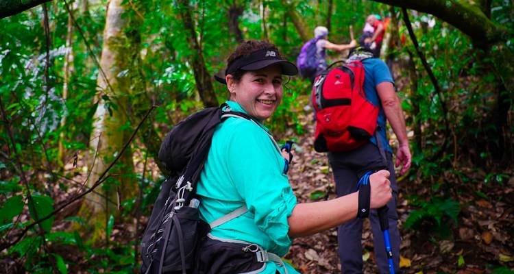 Lächelnder Wanderer mit Wanderstöcken auf einem blätterreichen Dschungelpfad, gefolgt von zwei Begleitern mit Rucksäcken.
