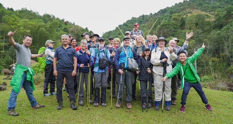 Große Gruppe von Wanderern posiert fröhlich mit Trekkingstöcken auf einem grasbewachsenen Hügel.
