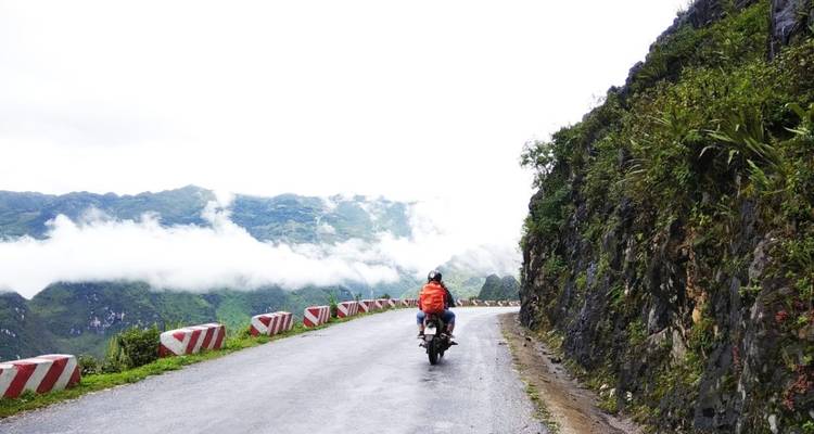 Ein einsamer Motorradfahrer fährt entlang einer Bergstraße, die von Wolken und felsigen Klippen gesäumt ist.