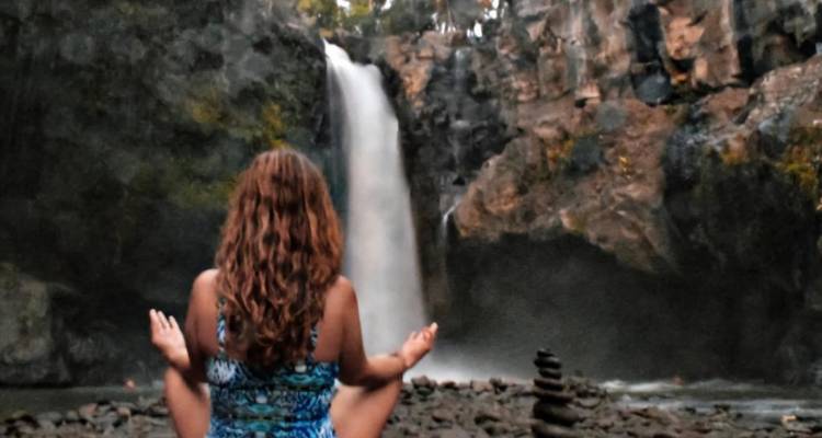 A person meditating in front of a waterfall with stacked stones.