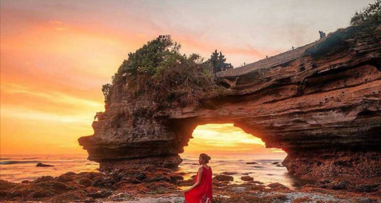 Person in a red dress sitting in front of a coastal rock arch at sunset.