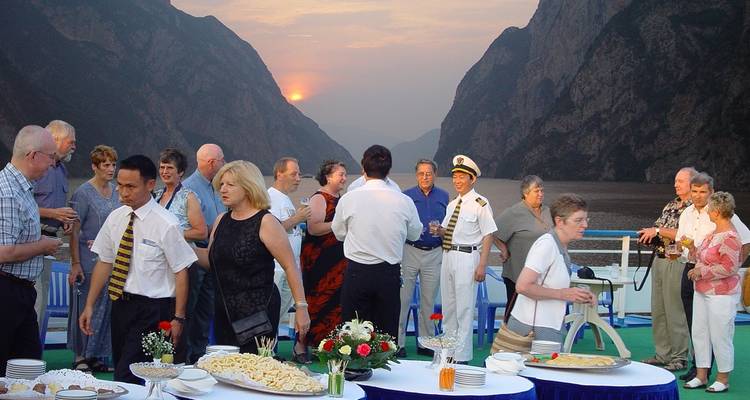 People socializing on a cruise ship deck at sunset in a mountain region.