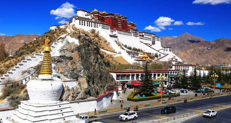 Breites Tagespanorama des Potala-Palastes, der sich über eine belebte Straße von Lhasa erhebt, und weiße Stupa unter tiefblauem Himmel.