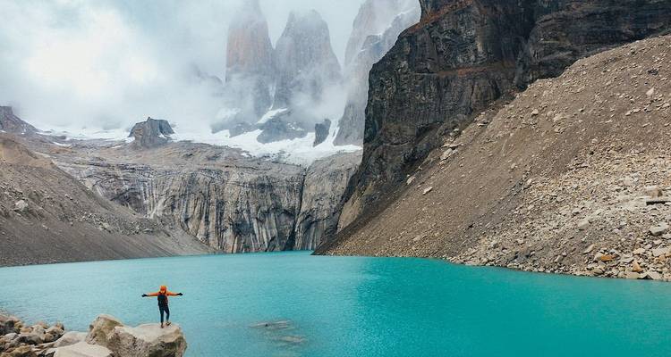 Person, die auf einem Felsen steht mit ausgebreiteten Armen vor einem atemberaubenden Bergsee.