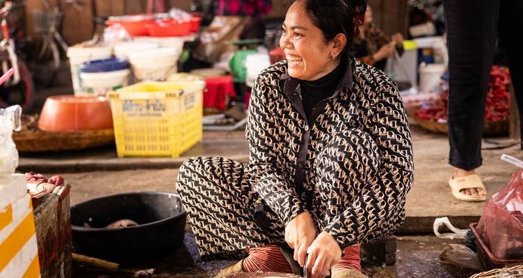 Un vendeur de marché souriant est assis par terre, entouré de paniers de produits.