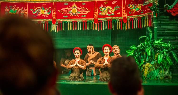 Traditionelle vietnamesische Wasserpuppenspieler begrüßen das Publikum mit gefalteten Händen auf einer grün beleuchteten Bühne.