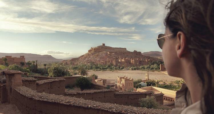 Seitenansicht eines Reisenden, der Ait Benhaddou und die trockene Landschaft von einer Lehmziegelmauer aus bewundert.