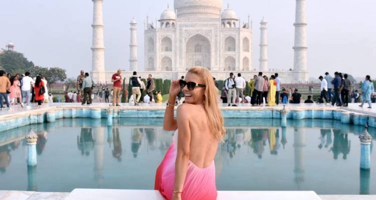 Femme élégante en robe rose dégradé posant près d'un bassin réfléchissant avec le Taj Mahal et la foule en arrière-plan.