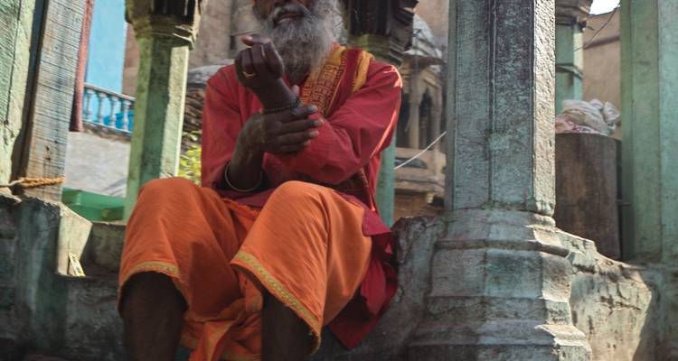 An elderly sadhu in orange robes sits and contemplates within a weathered stone pavilion.
