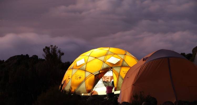 Illuminated tents under a cloudy night sky in a mountainous landscape.