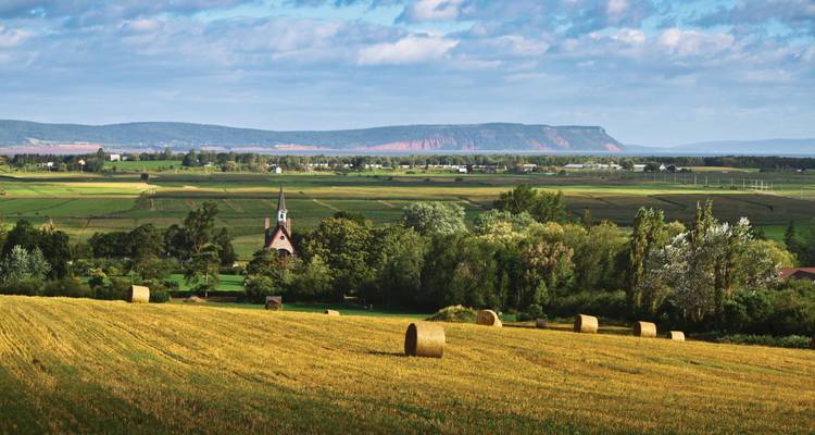 Terres agricoles ondulantes avec des balles de foin, un petit clocher d'église et des falaises lointaines sous un ciel aux motifs variés.
