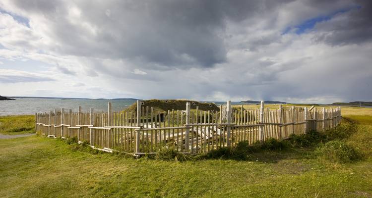 Un contour clôturé de maisons vikings reconstituées en tourbe se dresse sur un littoral herbeux sous des nuages spectaculaires.