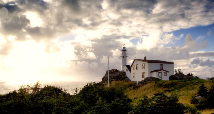 Un phare blanc et la maison du gardien se dressent au sommet d'un promontoire rocheux illuminés par une lumière dramatique en fin d'après-midi.