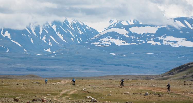 Fietsers rijden over een uitgestrekt stenig plateau in de richting van een besneeuwde bergketen onder lage wolken.