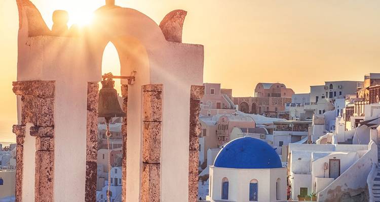 Sonnenuntergangsstrahlen scheinen durch einen weißen Glockenturm über Santorinis blau bekuppelten Dächern
