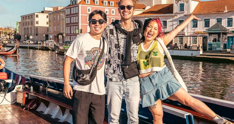 Tres viajeros posan junto a edificios coloridos y barcos tradicionales en el soleado frente de agua del canal de Aveiro.