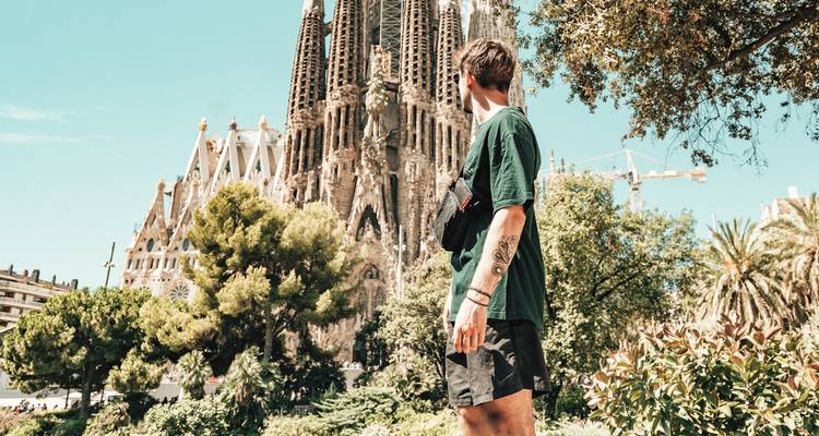 Un voyageur admire l'imposante Sagrada Família parmi la verdure luxuriante du parc sous un ciel dégagé.