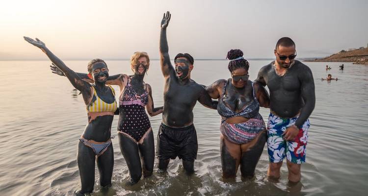 Des amis recouverts de boue noire riche en minéraux rient en se tenant debout dans la mer Morte jusqu'à la taille.