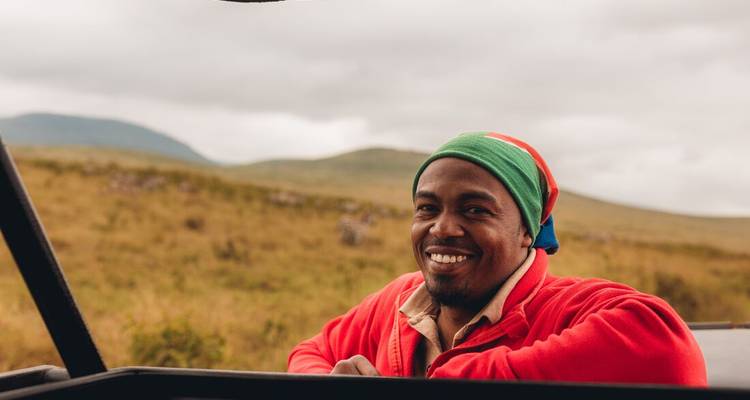 Un hombre sonriente en un vehículo de safari, llevando un sombrero colorido y rodeado de sabana.