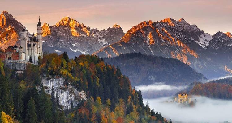 El Castillo de Neuschwanstein se alza sobre un acantilado boscoso frente a picos alpinos coronados de nieve que brillan con la luz del amanecer por encima de la niebla del valle