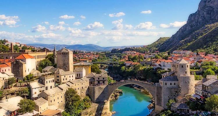 Un pont en arc de pierre traverse une rivière turquoise au milieu de la vieille ville historique de Mostar et des montagnes.