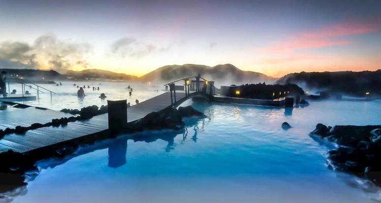 Hot spring with people in the water, surrounded by mist and sunset sky.