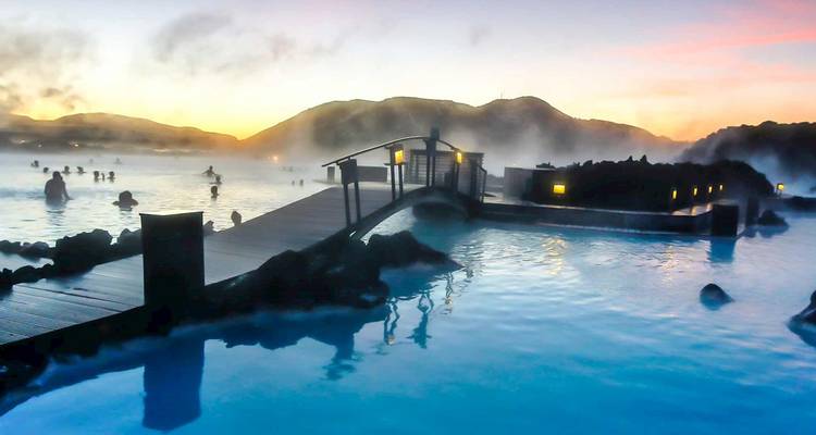 Piscines géothermales fumantes du Lagon Bleu au crépuscule avec des baigneurs se détendant près d'un pont en bois et des collines brumeuses au loin.