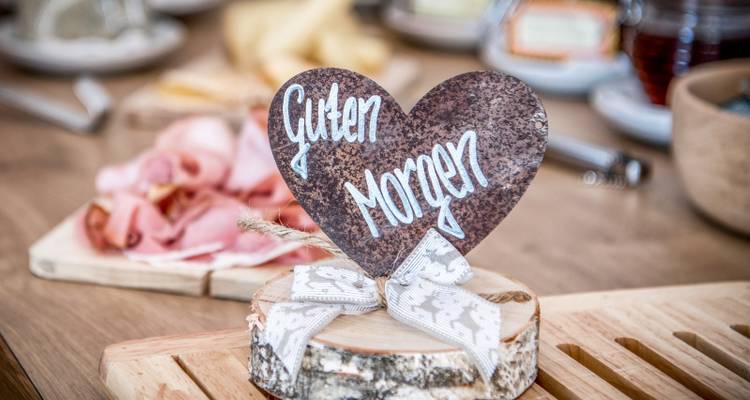 Table de petit-déjeuner rustique présentant un panneau en bois en forme de cœur inscrit « Guten Morgen » avec du jambon local en arrière-plan.