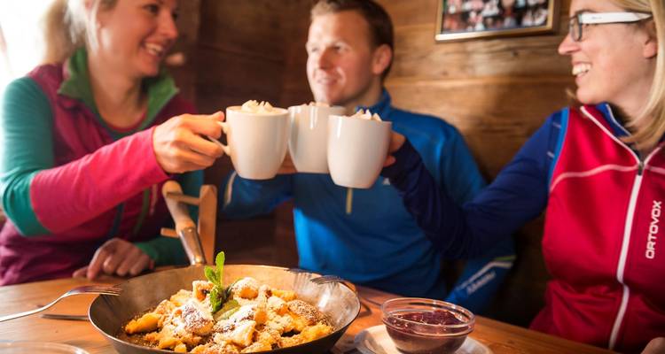 Trois amis vêtus de tenues de ski colorées trinquent avec des chopes au-dessus d'une poêle de cuisine alpine à l'intérieur d'une cabane en bois.
