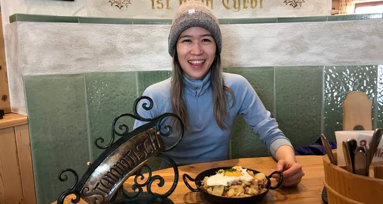 Femme souriante portant un bonnet d'hiver assise dans un restaurant alpin accueillant avec un repas traditionnel à la poêle.