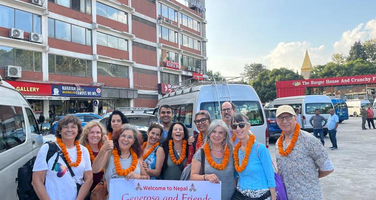 Un grand groupe de touristes portant des guirlandes de bienvenue sourit pour une photo dans un parking avec des vans et des bâtiments de la ville en arrière-plan.
