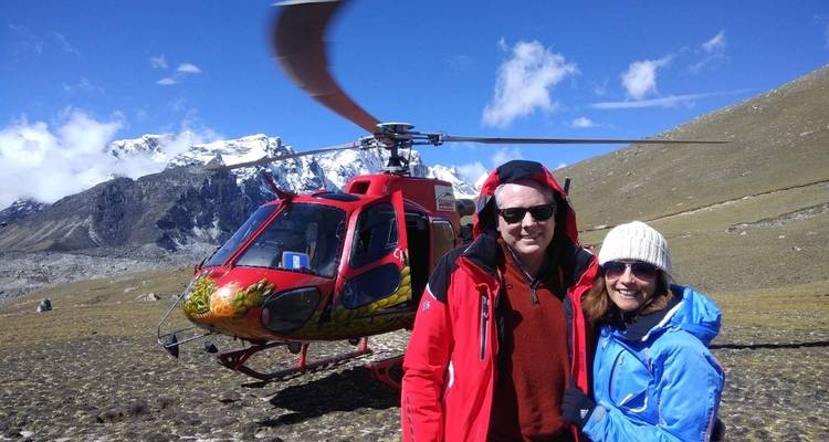 Zwei Personen posieren vor einem Hubschrauber mit Bergen im Hintergrund.