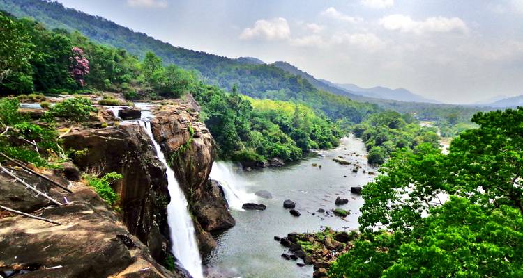 Una dramática cascada se desploma en un río rocoso rodeado de colinas verdes y montañas lejanas.