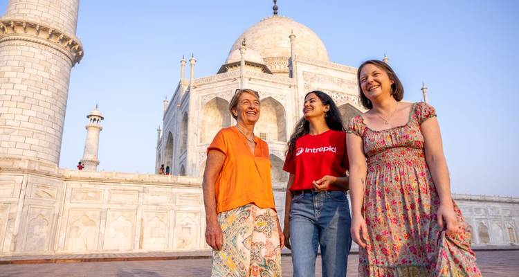 Tres mujeres ríen juntas frente al resplandeciente Taj Mahal de mármol blanco al amanecer.