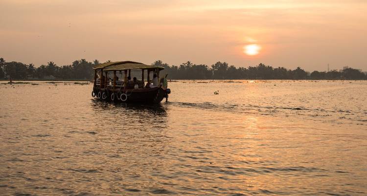 Un bateau en bois couvert glisse sur les paisibles backwaters du Kerala au coucher du soleil, le soleil orange se reflétant sur la large rivière.