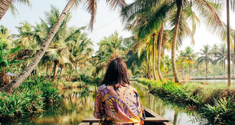 Une femme en robe à motifs est assise à la proue d'un canoë en bois qui dérive dans les canaux bordés de palmiers des backwaters du Kerala.