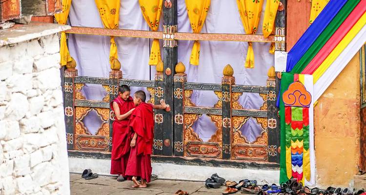 Zwei Mönche in roten Gewändern stehen vor einem verzierten Türeingang mit Schuhen auf dem Boden.