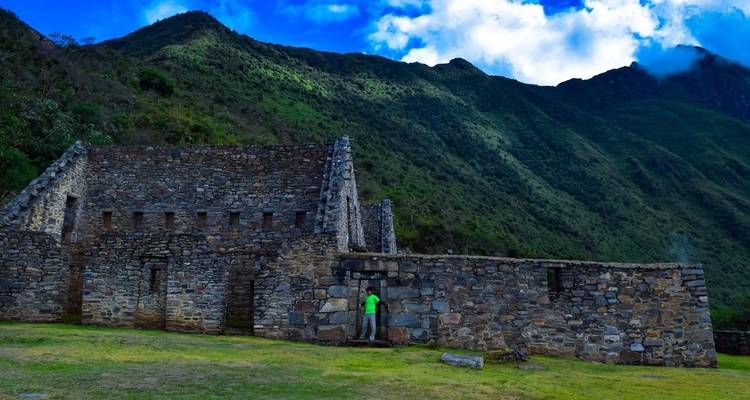 Person in a bright jacket near an ancient stone structure with lush green mountains in the background.