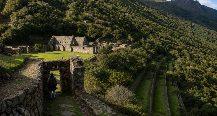 Stone gateway leads hikers toward extensive Incan-era ruins nestled in a forested mountain valley, lit by late-afternoon sun.