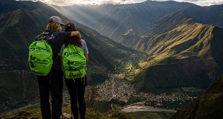 Zwei Wanderer mit passenden grünen Rucksäcken stehen Arm in Arm auf einem felsigen Aussichtspunkt und blicken auf ein sonniges Tal mit einer Stadt, die von steilen Andengipfeln umgeben ist.