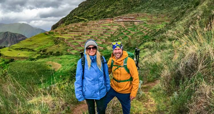 Smiling hikers pose on a mountain trail with sweeping agricultural terraces climbing the green hillside behind them under overcast skies.