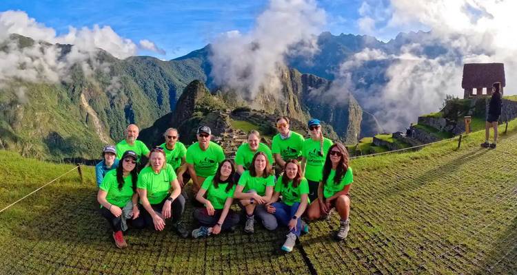 Tour group in matching neon shirts kneel and stand together on grassy terraces with the iconic ruins of Machu Picchu and misty peaks behind.