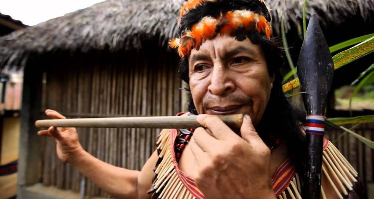 Un hombre indígena con un tocado de plumas toca una flauta de madera afuera de una choza con techo de paja.