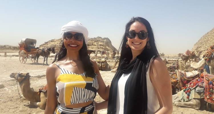 Deux femmes souriantes avec des pyramides et des chameaux en arrière-plan.