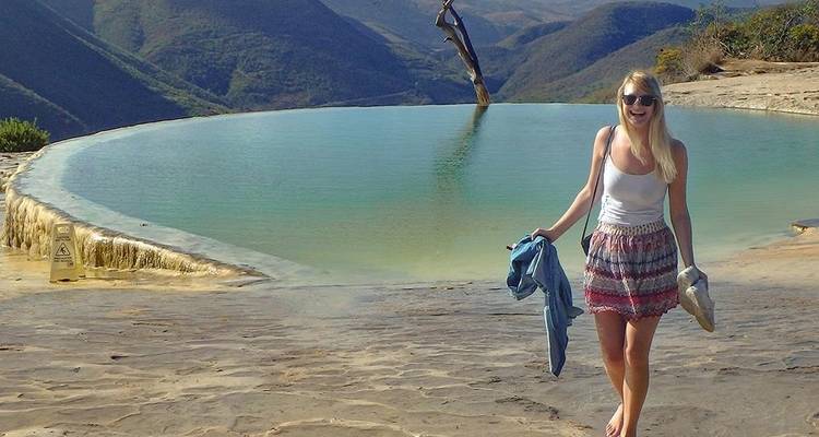 Une femme souriante se tient pieds nus à côté de la piscine à débordement naturelle de Hierve el Agua avec des montagnes en arrière-plan.