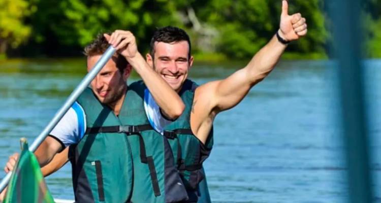 Zwei lächelnde Männer paddeln ein Boot auf einem Fluss, einer zeigt den Daumen nach oben.