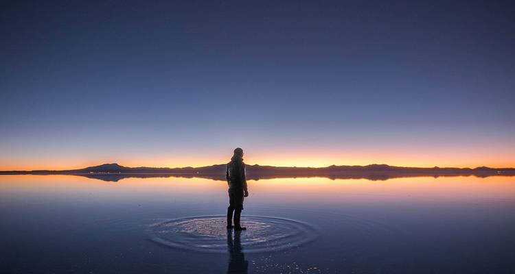 Een silhouet van een persoon die in ondiep water staat met een zonsondergang op de achtergrond in de Uyuni Zoutvlakte.