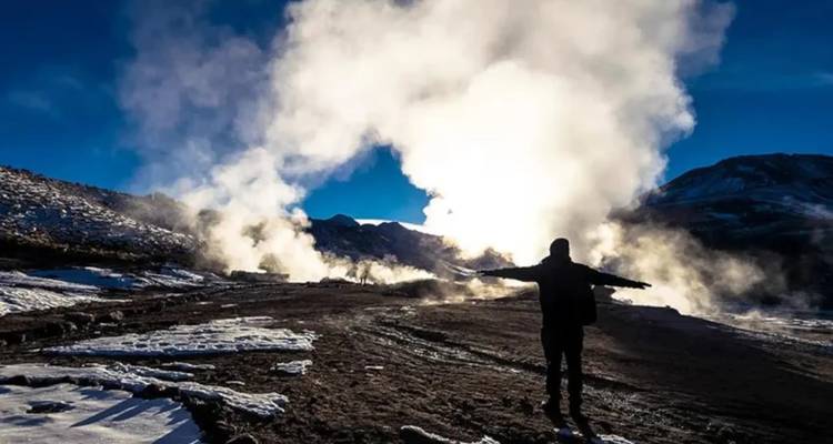 Una persona en silueta contra respiraderos de vapor y el amanecer en un terreno rocoso.